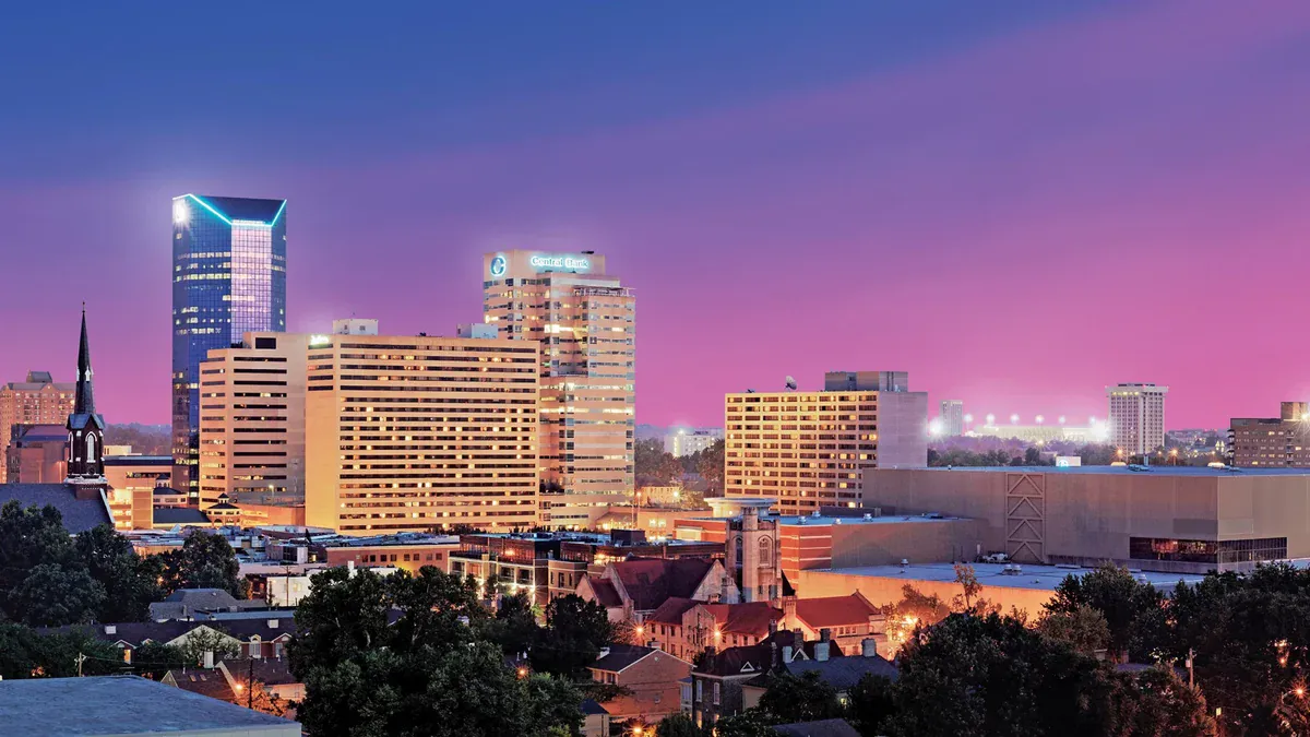 Illuminated downtown towers and church steeples rise above rooftops and trees beneath a purple and pink evening sky.
