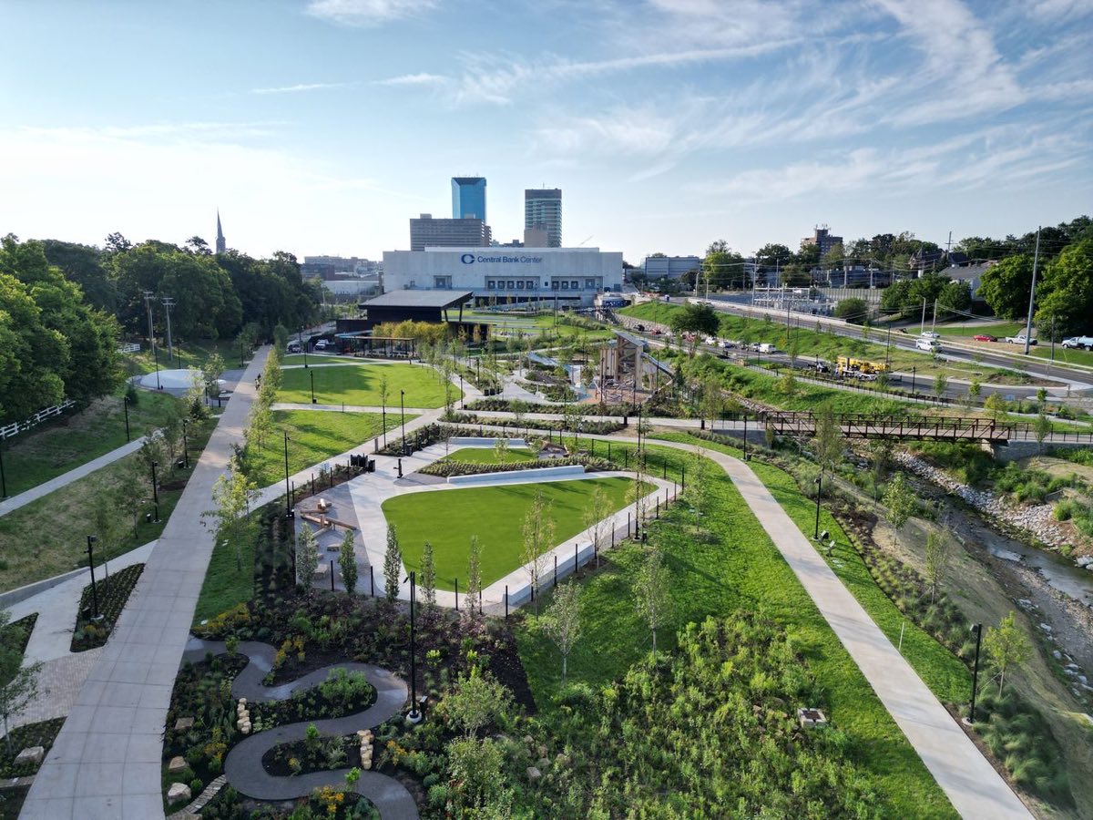 An elevated view of green lawns, winding paths, garden beds, and a footbridge over a creek, with downtown buildings beyond.