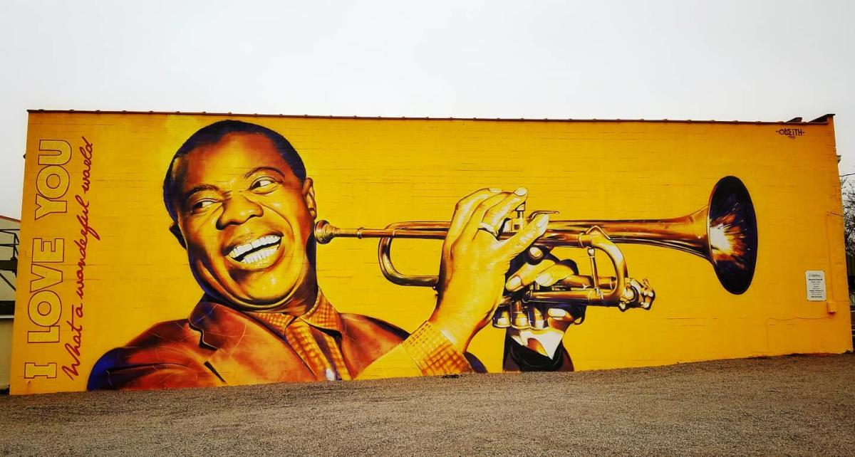 Golden-yellow mural of a smiling musician holding a trumpet across a building wall, beside the words ‘I LOVE YOU’ and ‘What a wonderful world.’